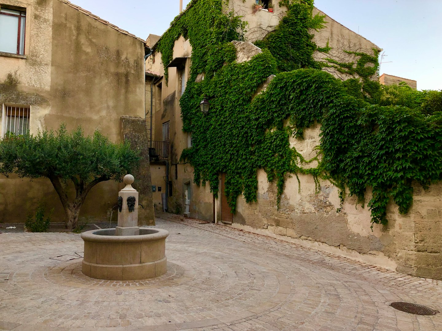 Fontana in pietra con volti scolpiti in una piazza soleggiata di un villaggio europeo.